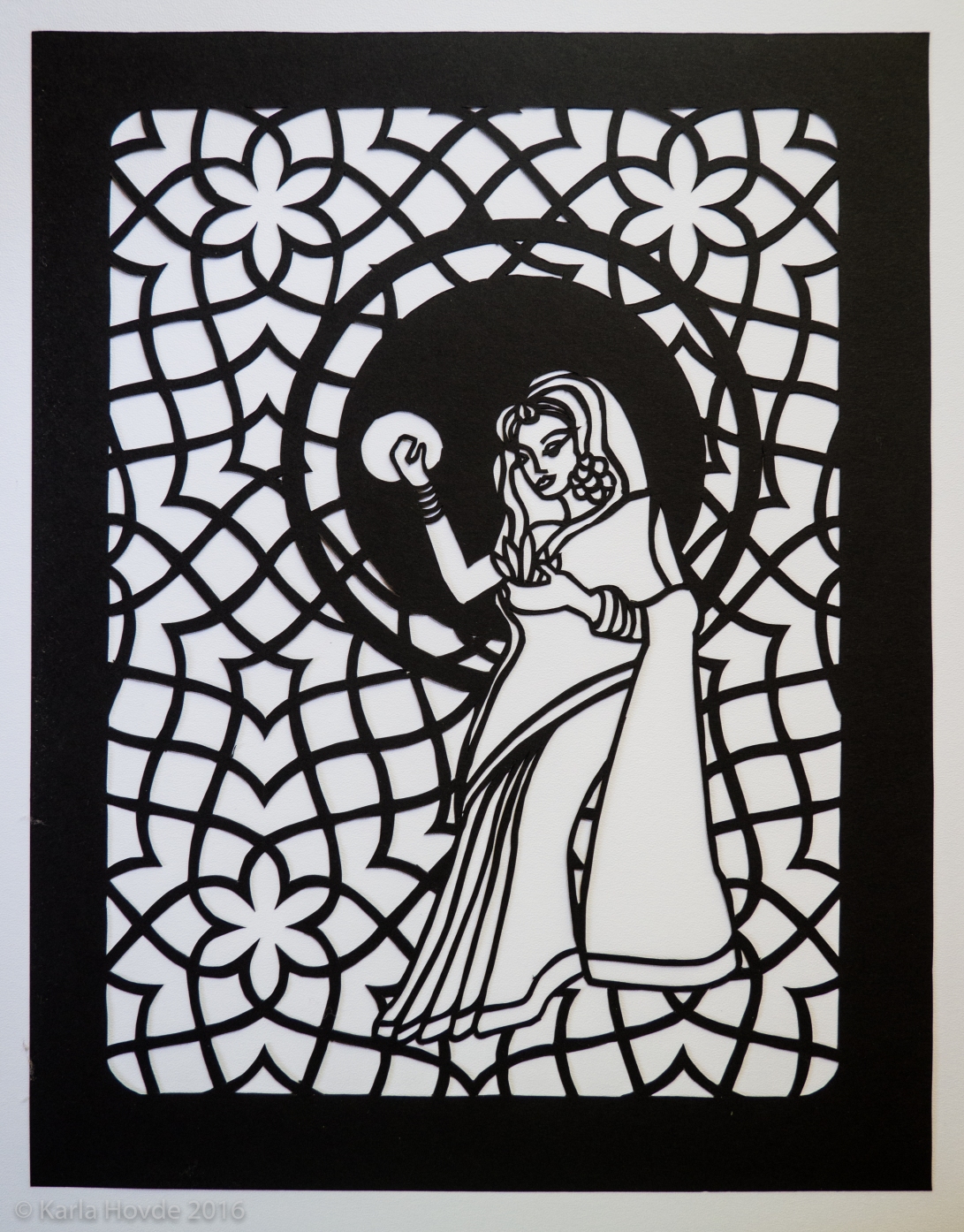 Paper cutting art in black paper shows stylized woman wearing a sari in front of delicate geometric arabesque pattern.